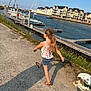 blue_sky, boats, buildings, casual_clothing, child, concrete, dock, fence, flowers, girl, harbor, marina, outdoor, pony_tail, sandals, shadow, skipping, summer, sunny, water