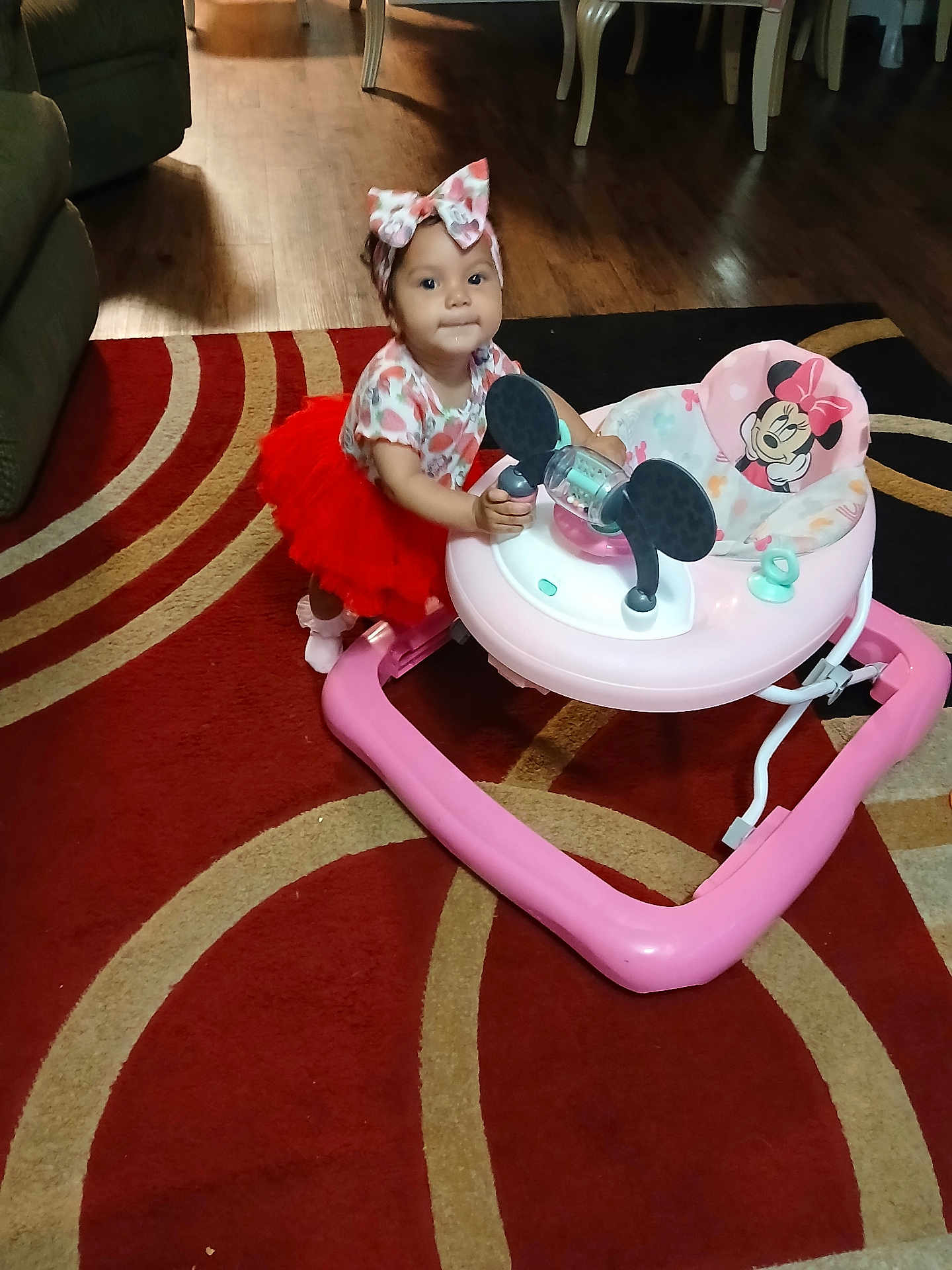 Marcelina is registered to the contest to win money with this photo: toddler, baby, child, walker, red_tutu, headband, indoor, carpet, flooring, living_room, toy, pink, mickey_mouse, furniture, person, cute, smile, playtime, floor, curious