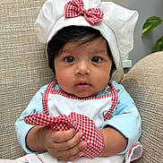 Amaira is registered to the contest to win money with this photo: baby, child, chef_hat, apron, gingham, red_bow, cute, curious_eyes, indoor, seated, hand, clothing, portrait, furniture, beige_chair, soft_light, infant, face, expression, holding