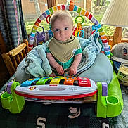 Kaleb joined the competition — help win amazing prizes! baby, infant, toy_piano, activity_seat, blanket, bib, curious, indoor, window, wooden_chair, lamp, hat, candle, table, green, colorful, face, person, child, playtime