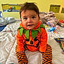 baby, bed, blanket, child, costume, curious, cute, face, green, halloween, holiday, indoors, orange, person, plush_toy, pumpkin_costume, sitting, smile, striped_pants, toy