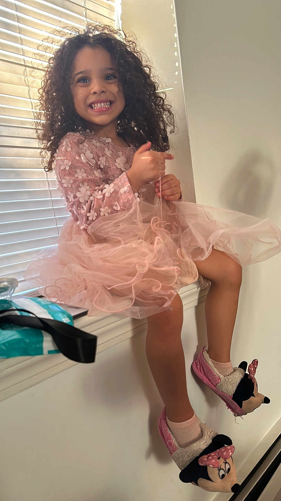 Pietra is registered to the contest to win money with this photo: child, girl, smile, curly_hair, pink_dress, sitting, window, blinds, natural_light, slippers, minnie_mouse, happy, indoor, legs, playful, cute, cozy, home, fashion, portrait