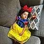 child, toddler, sleeping, dress, costume, snow_white, red_bow, apple, pillow, couch, gray_couch, curly_hair, indoors, relaxation, rest, fairy_tale, yellow_skirt, blue_top, cute, peaceful