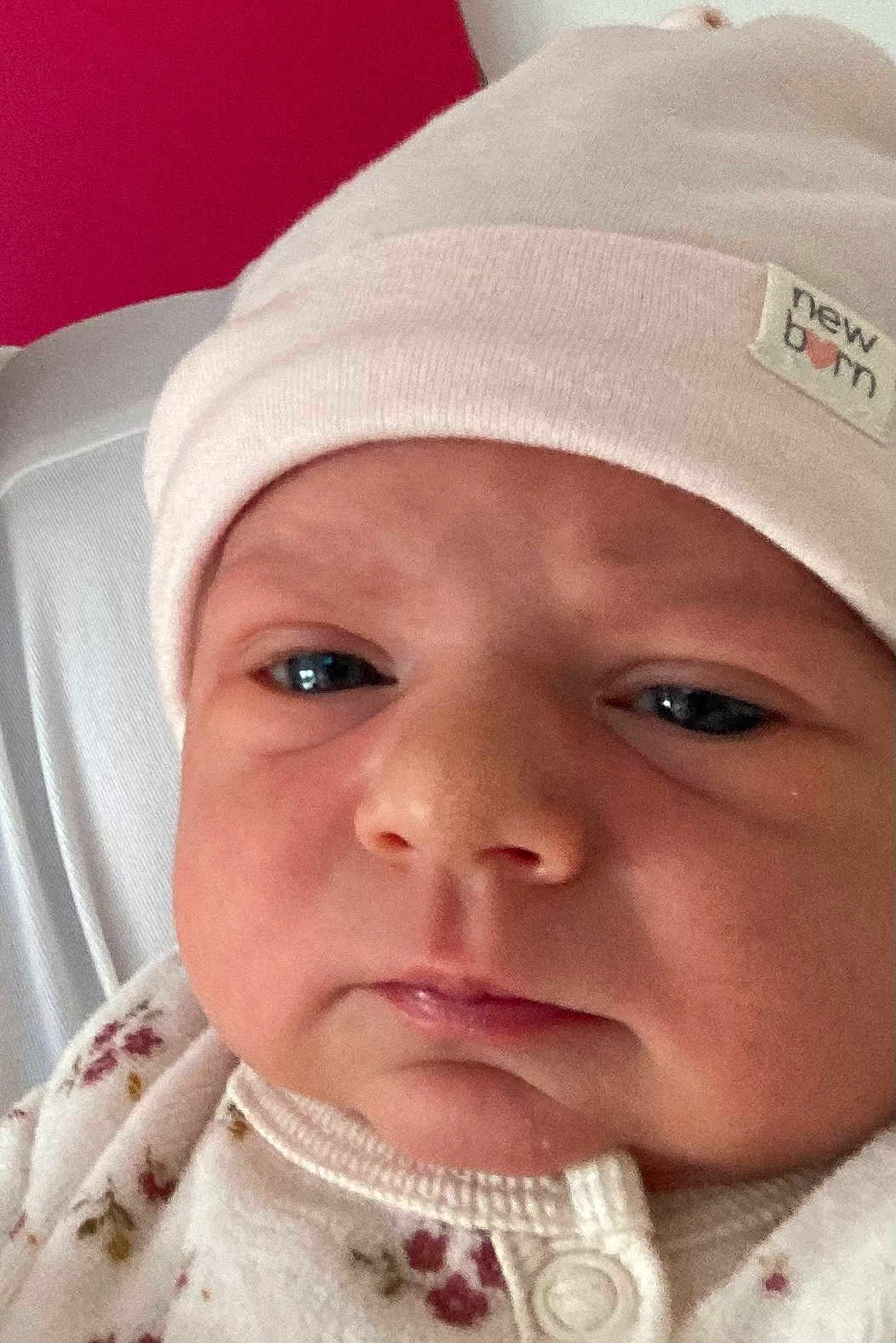Justine participe au concours pour gagner de l'argent avec cette photo : baby, newborn, hat, face, closeup, expression, clothing, pink, white, red_background, infant, skin, eyes, mouth, nose, soft_fabric, button, sleepwear, person, portrait