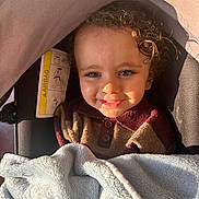 Ali a rejoint le concours — aidez-le/la à gagner de superbes lots ! toddler, child, smile, curly_hair, blanket, stroller, sunlight, shadow, face, clothing, buttoned_shirt, cozy, warm, baby, portrait, indoor, happy, person, cute, infant