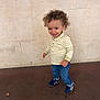 toddler, child, curly_hair, striped_shirt, polo_shirt, blue_jeans, sneakers, standing, smiling, happy, indoor, wall, floor, person, young_child, casual_clothing, portrait, full_body, light_skin, cute