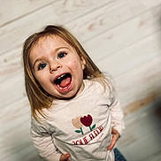 Laïss a rejoint le concours — aidez-le/la à gagner de superbes lots ! background_blur, blonde_hair, blue_eyes, child, close_up, earrings, floral_graphic, girl, happy, indoor, long_hair, natural_light, open_mouth, patterned_pants, playful, portrait, shirt, smile, toddler, wooden_wall