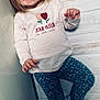 Laïss participe au concours pour gagner de l'argent avec cette photo : blonde_hair, blue_eyes, casual_clothing, child, cute, floral_leggings, graphic_print, hands, indoor, leaning, one_face, portrait, smiling, sneakers, standing, teeth, toddler, white_shirt, wooden_background, young