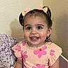 Sariyah is registered to the contest to win money with this photo: toddler, child, girl, smiling, happy, indoor, hair_bows, pink_flower_shirt, denim_skirt, sitting, face, eyes, cute, clothing, blanket, pillow, wall, portrait, person, young_child