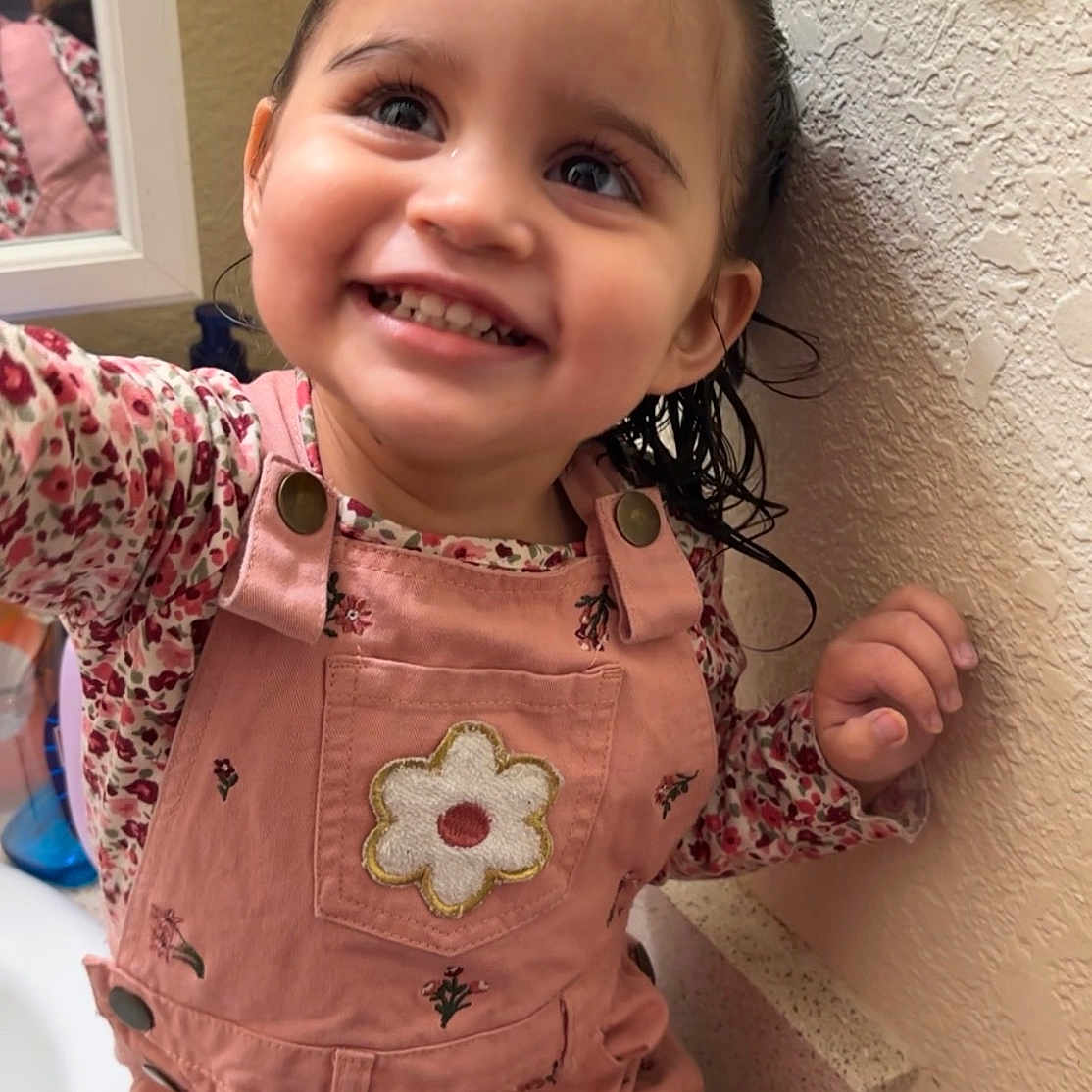 Sariyah is registered to the contest to win money with this photo: toddler, child, smiling, pink_overalls, floral_pattern, wet_hair, bathroom, sink, mirror, wall, portrait, happy, cute, indoor, person, young_child, hand, face, clothing, countertop