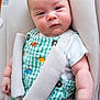 baby, infant, car_seat, safety_harness, green_overalls, dinosaur_pattern, white_shirt, cute, chubby_cheeks, face, eyes, arm, hand, fabric_texture, button, child, portrait, indoors, seat_belt, soft_lighting