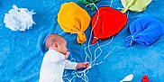 James is registered to the contest to win money with this photo: baby, blue_blanket, colorful_balloons, cotton_clouds, fabric_balloons, infant, lying_down, toy_birds, white_clothing, white_socks, strings, soft_texture, playful_scene, hand, head, peaceful, child, indoor, creative, artistic
