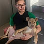 man, dog, glasses, indoor, stairs, sitting, casual, relaxed, brown_dog, black_pants, green_shirt, floor, door, pet, holding, portrait, young_adult, home, curious, calm
