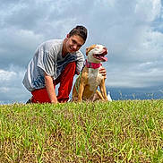 Buffy is registered to the contest to win money with this photo: man, dog, grass, outdoor, sky, clouds, smiling, collar, pet, happy, casual_clothing, nature, daylight, greenery, playing, friendship, animal, person, crouching, tongue_out