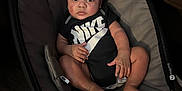 Jaylen is registered to the contest to win money with this photo: baby, infant, child, nike_onesie, baby_rocker, cushion, indoor, carpet, brown_couch, footwear, curly_hair, looking_at_camera, relaxed, seated, young_child, person, clothing, furniture, floor, expression