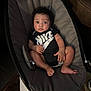 baby, infant, child, nike_onesie, baby_rocker, cushion, indoor, carpet, brown_couch, footwear, curly_hair, looking_at_camera, relaxed, seated, young_child, person, clothing, furniture, floor, expression