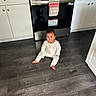 baby, pajamas, kitchen, floor, oven, cabinets, stainless_steel, smile, child, indoor, appliance, towel, wood_floor, person, home, footwearless, cabinet_knobs, curly_hair, infant, cute