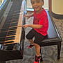 child, piano, bench, indoor, carpet, window, smiling, red_shirt, black_shorts, sneakers, musical_instrument, happy, person, fingers, seated, young_boy, light, reflection, floor, casual_clothing