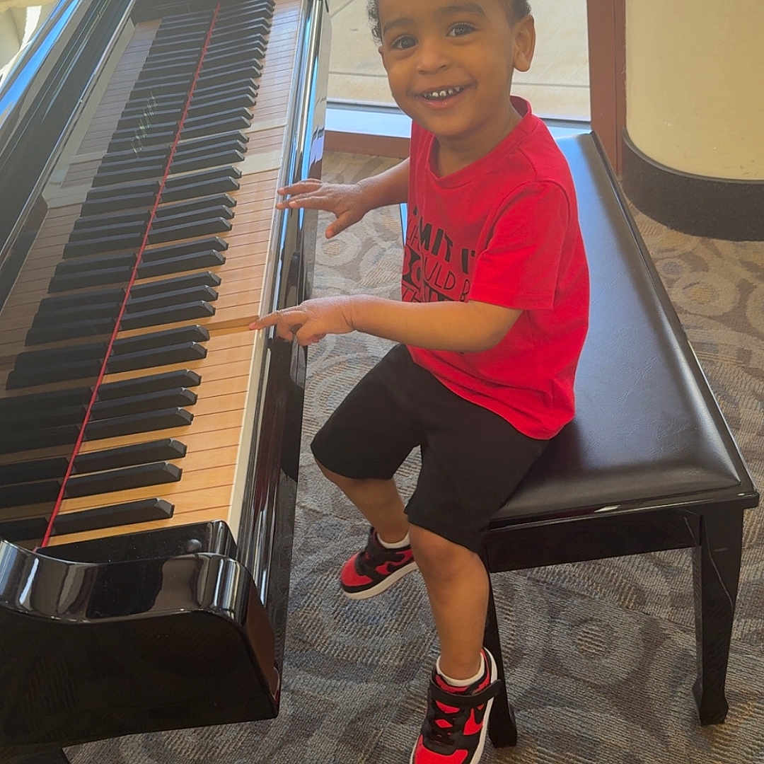 Ladedrick joined the competition — help win amazing prizes! bench, black_shorts, carpet, casual_clothing, child, fingers, floor, happy, indoor, light, musical_instrument, person, piano, red_shirt, reflection, seated, smiling, sneakers, window, young_boy