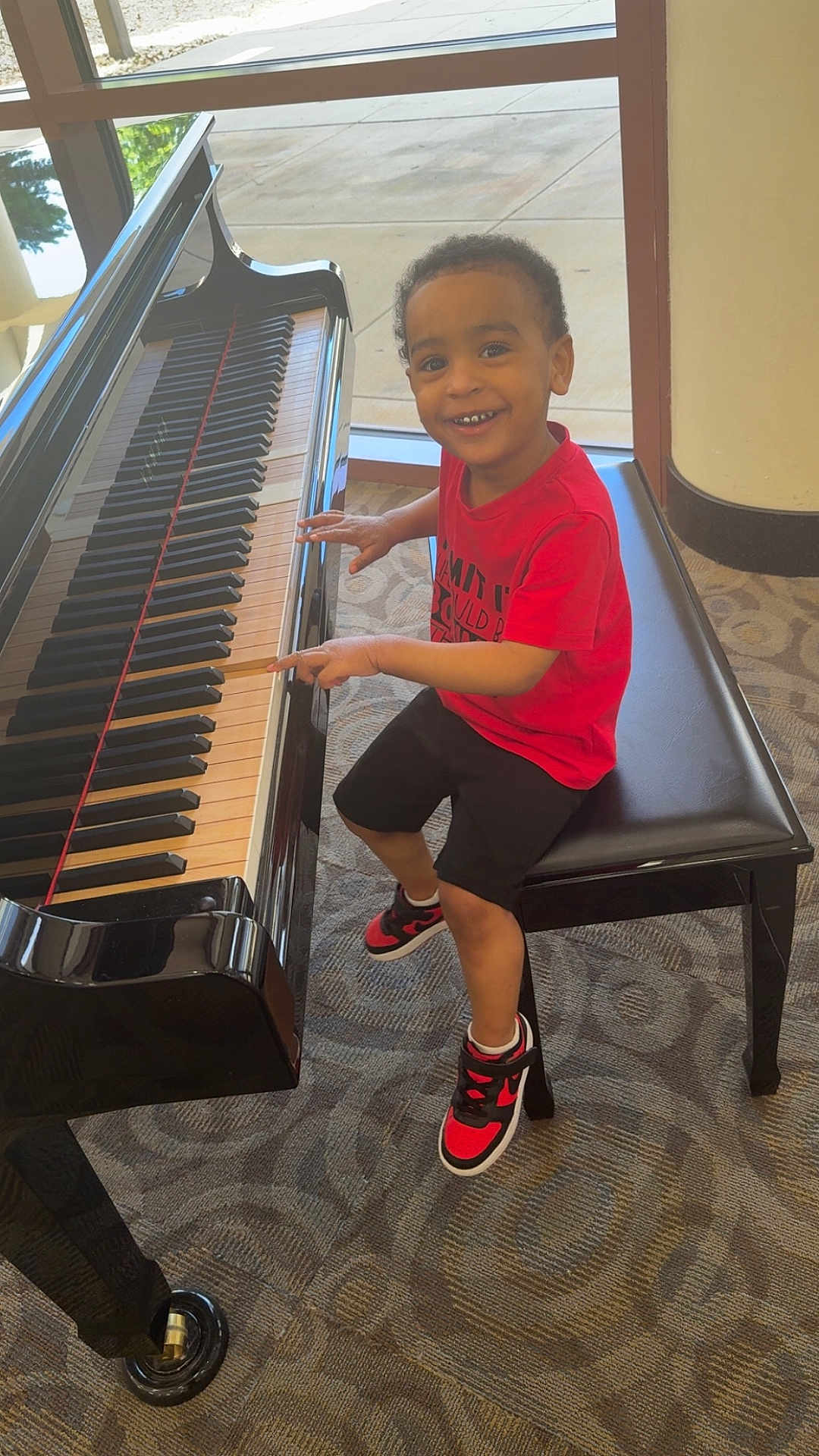 Ladedrick joined the competition — help win amazing prizes! child, piano, bench, indoor, carpet, window, smiling, red_shirt, black_shorts, sneakers, musical_instrument, happy, person, fingers, seated, young_boy, light, reflection, floor, casual_clothing