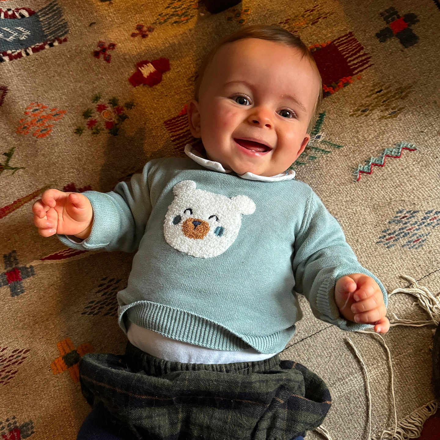 Arnould participe au concours pour gagner de l'argent avec cette photo : baby, bear_design, blue_tights, child, clothing, cozy, cute, face, floor, hands, happy, indoor, infant, lying_down, patterned_rug, person, playing, rug, smiling, sweater