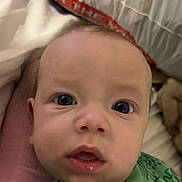 Hayden is registered to the contest to win money with this photo: baby, infant, face, close_up, green_clothing, dinosaur_print, bed, pillow, curious, wide_eyes, mouth_open, sleepwear, child, person, indoors, soft_lighting, skin, head, young, portrait