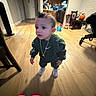 Luis is registered to the contest to win money with this photo: toddler, child, indoors, home, standing, portrait, face, curious, clothing, sneakers, green_outfit, necklace, wooden_floor, kitchen, living_room, chair, desk_chair, toy, pumpkin, heart