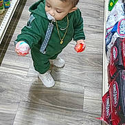 Luis is registered to the contest to win money with this photo: child, toddler, baby, supermarket, grocery_store, shopping_aisle, green_tracksuit, pacifier, gold_chain, candy, lollipop, holding_objects, standing, sneakers, floor, shelves, packaged_goods, portrait, indoors, funny_pose