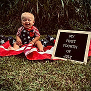 Teegan joined the competition — help win amazing prizes! baby, child, grass, american_flag, patriotic, smiling, outdoor, sign, celebration, holiday, clothing, summer, nature, greenery, happy, portrait, sitting, cute, red_white_blue, infant