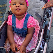 Rhythm is registered to the contest to win money with this photo: toddler, child, stroller, pink, denim_overalls, boots, outdoor, person, hands, hair_beads, clothing, footwear, seated, casual, daylight, urban, cute, baby, fashion, street