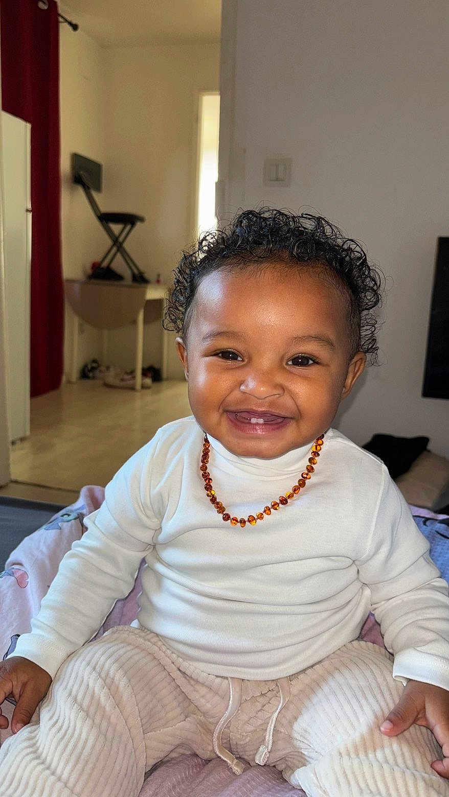 Aneïa a rejoint le concours — aidez-le/la à gagner de superbes lots ! toddler, child, smiling, curly_hair, white_shirt, beige_pants, necklace, indoor, bed, blanket, happy, person, home, casual, floor, furniture, curtain, light, portrait, cute