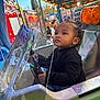 amusement_park, black_shirt, carnival, casual_clothing, child, colorful, curious, daytime, fun, hair, lights, outdoor, playful, portrait, ride, seat, steering_wheel, toddler, vehicle, young_child
