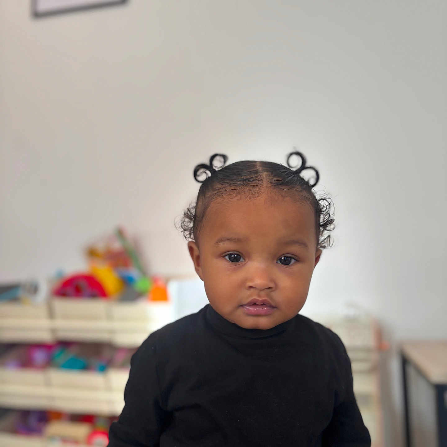 Aneïa participe au concours pour gagner de l'argent avec cette photo : black_clothing, blurred_background, casual, child, colorful, curly_hair, expression, eyes, face, furniture, hair_buns, home, indoor, person, portrait, shelf, table, toddler, toys, young_child