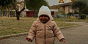 Aneïa a rejoint le concours — aidez-le/la à gagner de superbes lots ! black_shoes, building, child, cold_weather, daytime, fence, grass, jeans, knit_hat, nature, outdoor, overcast_sky, park, pathway, person, puffer_jacket, smiling, toddler, trees, walking