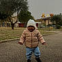 black_shoes, building, child, cold_weather, daytime, fence, grass, jeans, knit_hat, nature, outdoor, overcast_sky, park, pathway, person, puffer_jacket, smiling, toddler, trees, walking