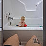 bath_time, bathroom, bathtub, blond_hair, blue_toy, child, faucet, home, indoor, legs, phone_screenshot, portrait, reaction_icons, red_ball, sitting, tattoo, toddler, toy, water, white_tub