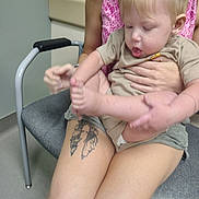 Keagan is registered to the contest to win money with this photo: toddler, child, adult, tattoo, chair, foot, hand, shorts, shirt, blond_hair, indoor, person, seat, curious, skin, cushion, floor, wall, casual, interaction