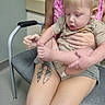 toddler, child, adult, tattoo, chair, foot, hand, shorts, shirt, blond_hair, indoor, person, seat, curious, skin, cushion, floor, wall, casual, interaction