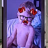  스마트phone, baby, bed, bedding, bedroom, blanket, cartoon_filter, child, diaper, face_filter, glasses, horns, humorous, indoor, infant, mustache, portrait, purple_lighting, screenshot, sitting
