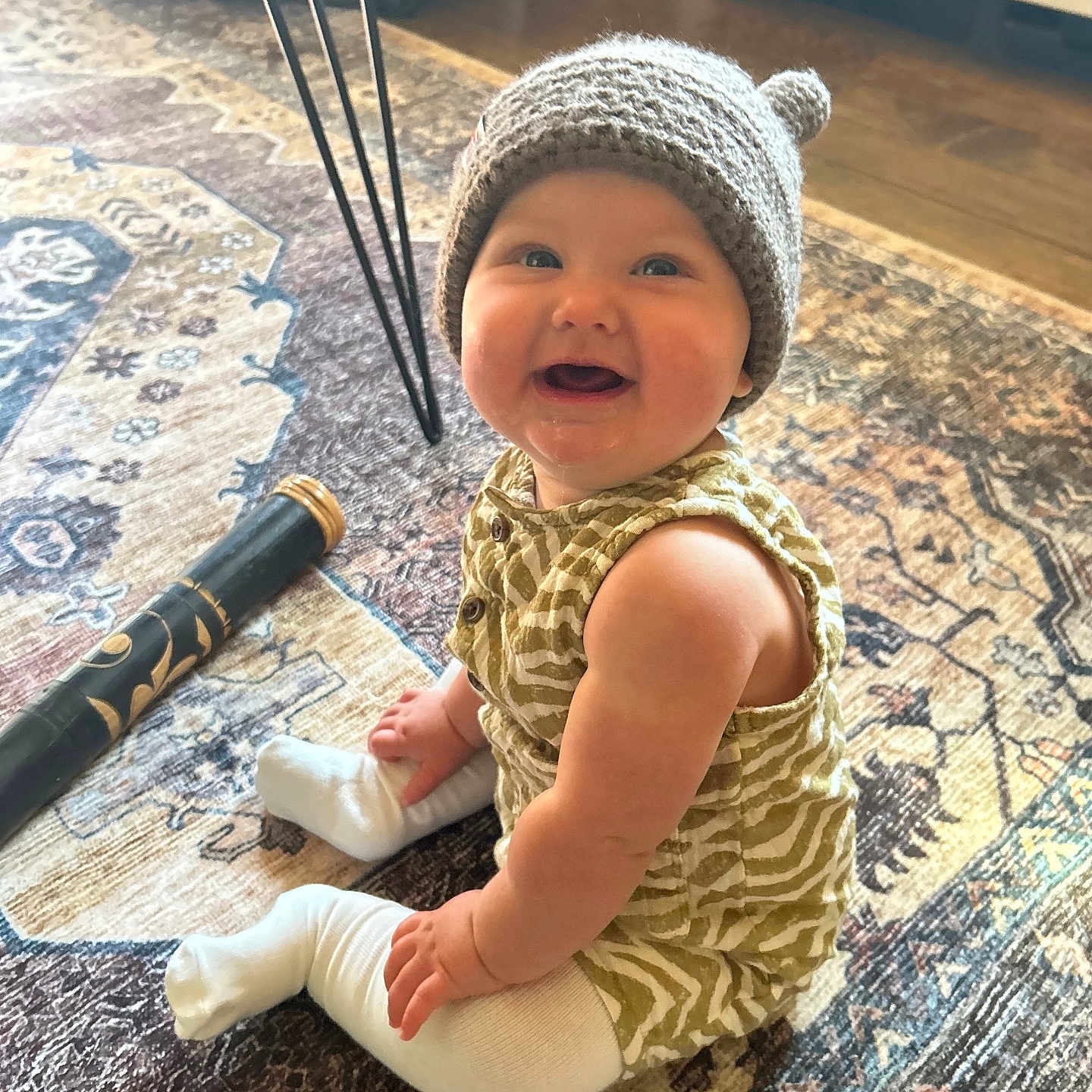 Tulsi joined the competition — help win amazing prizes! baby, carpet, chubby_cheeks, foot, hand, happy, hardwood_floor, hat, indoor, infant, knit_hat, patterned_rug, portrait, romper, sitting, smile, socks, table_leg, toy, white_cabinet