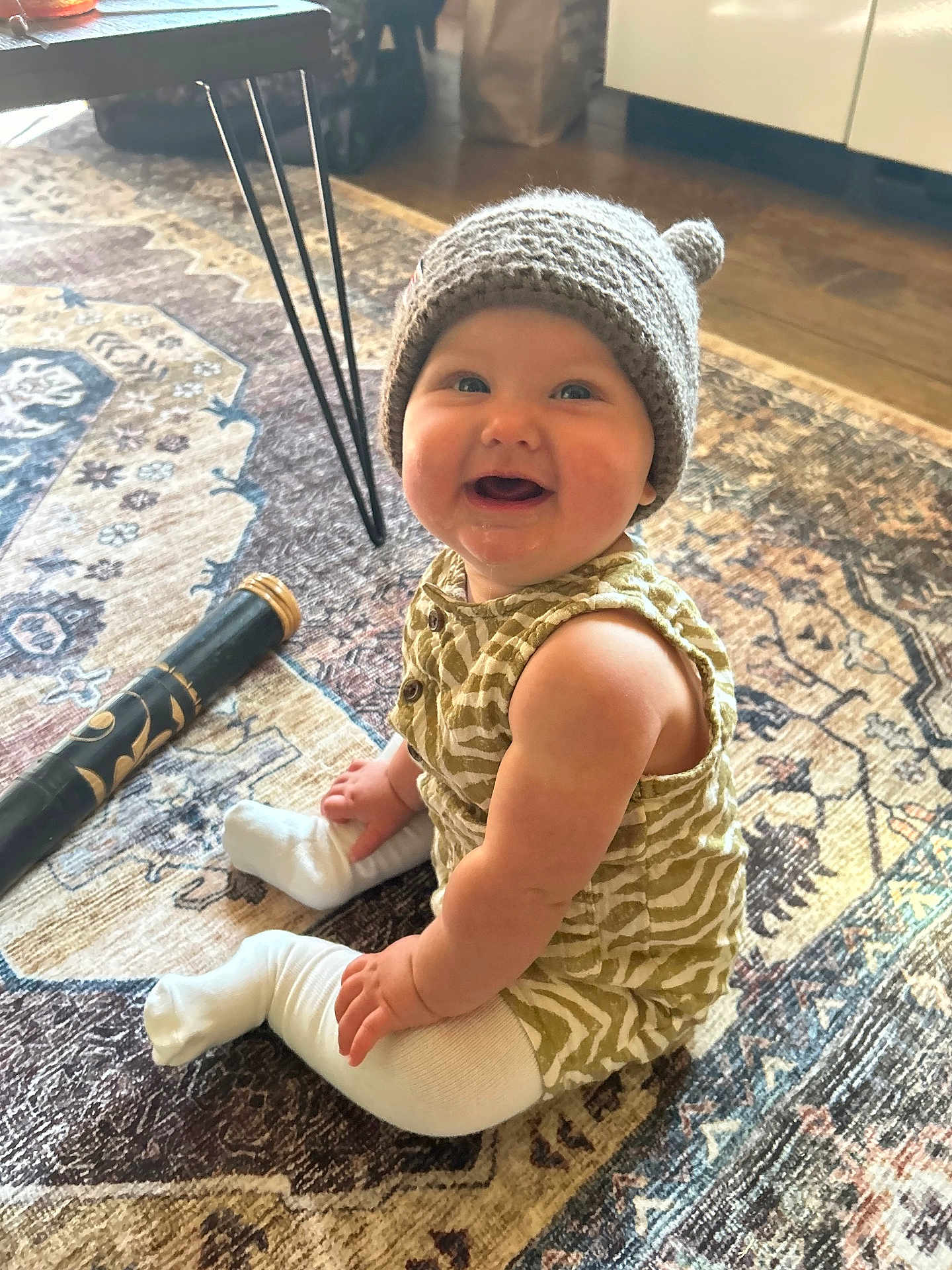 Tulsi joined the competition — help win amazing prizes! baby, infant, smile, knit_hat, hat, sitting, patterned_rug, carpet, indoor, hardwood_floor, table_leg, white_cabinet, romper, socks, chubby_cheeks, happy, portrait, toy, hand, foot