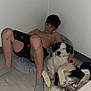 boy, dog, bean_bag, nintendo_switch, indoor, floor, casual, relaxed, pet, companion, shorts, socks, light_skin, black_hair, white_wall, wooden_floor, playing, technology, comfort, resting