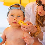 Edgard a rejoint le concours — aidez-le/la à gagner de superbes lots ! toddler, child, adult, beach, sunscreen, umbrella, hat, blue_eyes, striped_shorts, summer, sunlight, outdoor, skin_care, toy, casual, bare_chest, protective, care, family, vacation