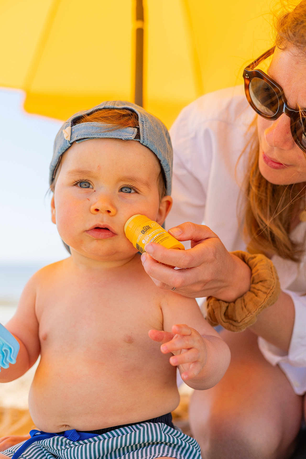 Edgard a rejoint le concours — aidez-le/la à gagner de superbes lots ! toddler, child, adult, beach, sunscreen, umbrella, hat, blue_eyes, striped_shorts, summer, sunlight, outdoor, skin_care, toy, casual, bare_chest, protective, care, family, vacation