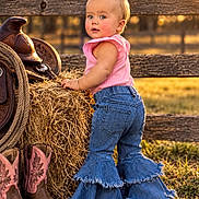 Callie is registered to the contest to win money with this photo: child, toddler, cowgirl, outdoor, portrait, smiling, blonde_hair, hair_bow, pink_shirt, denim, jeans, bell_bottoms, cowboy_boots, saddle, hay_bale, fence, farm, ranch, sunset, grass