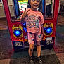 child, arcade, game, peace_sign, smiling, pink_clothing, shorts, tshirt, indoor, carpet, sneakers, standing, person, gaming_machine, colorful, blue_light, happy, casual, young, fun