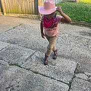 Malaysia is registered to the contest to win money with this photo: child, cowboy_hat, bandana, boots, outdoor, gravel, concrete, grass, fence, sidewalk, posing, pink_clothing, braids, glasses, casual_wear, sunlight, young, person, footwear, costume