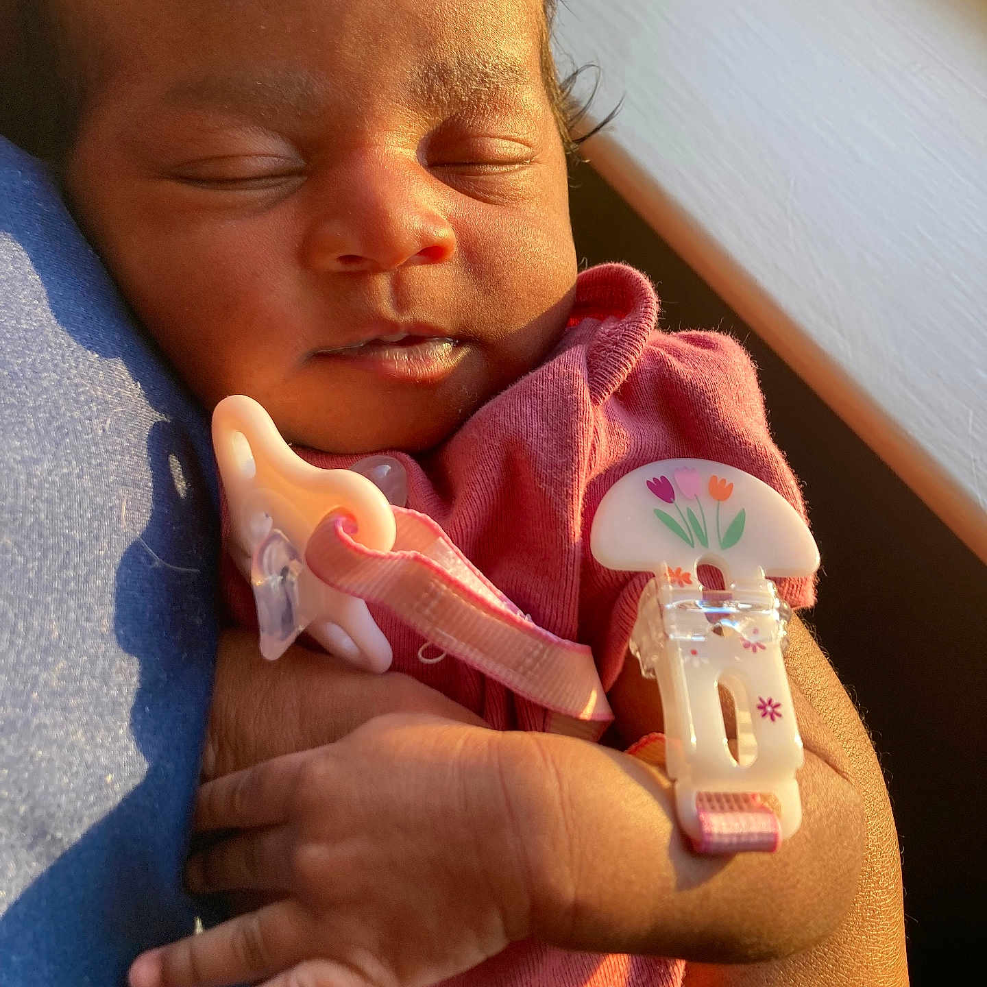 Dream is registered to the contest to win money with this photo: newborn, baby, sleeping, infant, portrait, pink_clothing, pacifier, pacifier_clip, floral_design, adult_arm, cuddling, close_up, warm_lighting, sunlight, indoor, cozy, peaceful, child, face, hands