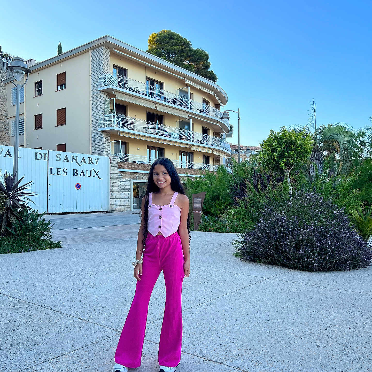 Ana Bheatriz a rejoint le concours — aidez-le/la à gagner de superbes lots ! building, casual_clothing, child, concrete, daytime, girl, greenery, happy, long_hair, outdoor, pants, person, plants, sky, smiling, sneakers, standing, top, tree, urban