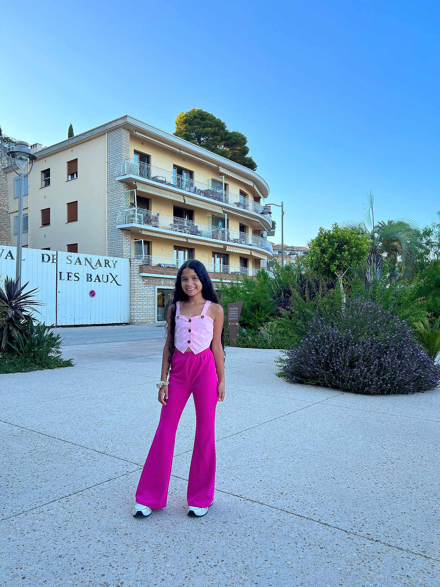 Ana Bheatriz a rejoint le concours — aidez-le/la à gagner de superbes lots ! child, girl, smiling, standing, outdoor, building, greenery, tree, sky, casual_clothing, long_hair, pants, top, sneakers, urban, daytime, plants, concrete, happy, person